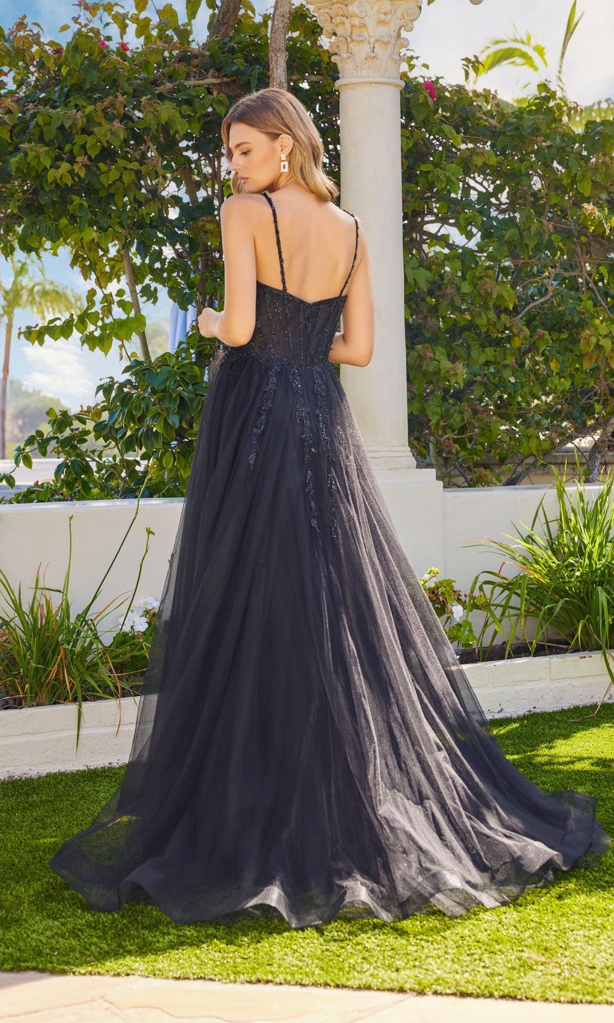 A woman stands outdoors in the Juliet JT2632L Long Prom Dress, featuring a flowing tulle A-line skirt and thin straps. Surrounded by lush greenery and stone columns, her elegant silhouette is seen from behind.